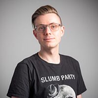 A colour photo of a young white man, the artist James Medd. He wears glasses with his hair swept back from his face. He wears a black t-shirt adorned with the words 'Slumber Party' and an illustration of the moon wearing a top hat.