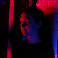 A colour photograph of a young white woman, the artist Izzy Bolt. She is cast in dramatic pink and blue lighting, and lots of shadow. She looks out of frame to the left, smiling.