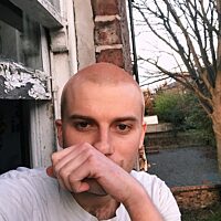 A colour photograph of a young white man, the artist Jacob Bolton. His hair is shaved and he wears aa white t-shirt, he looks into camera while his left hand rests just in front of his chin