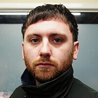 A colour photograph of a young white man, the artist Joseph Whitmore, looking directly into the camera. He has cropped brown hair and beard, and is wearing a dark jacket with a high neck.
