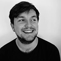A black and white photograph of young white man, the artist David McFarlane, smiling and looking just out of frame. He is wearing a black t-shirt